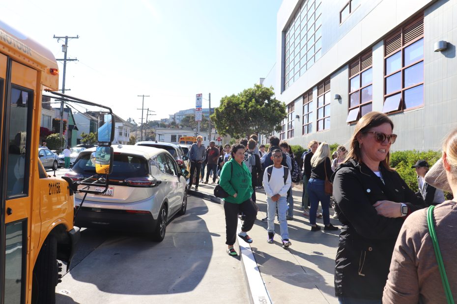 A group of people, including students, stands and walks on a sidewalk near parked cars and a school bus outside a modern building on a sunny day.