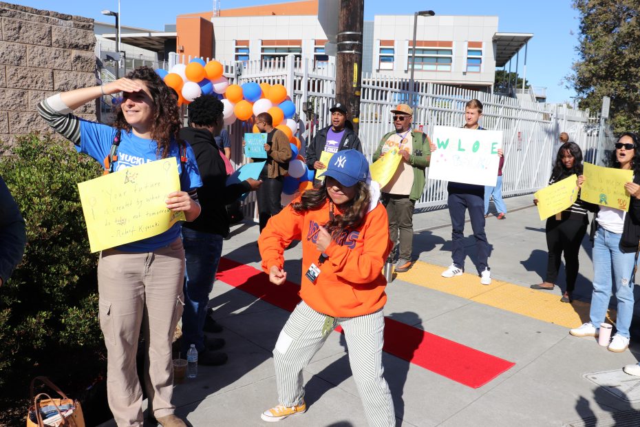 People stand outdoors holding colorful signs and balloons near a building; one person dances while others watch or look into the distance.