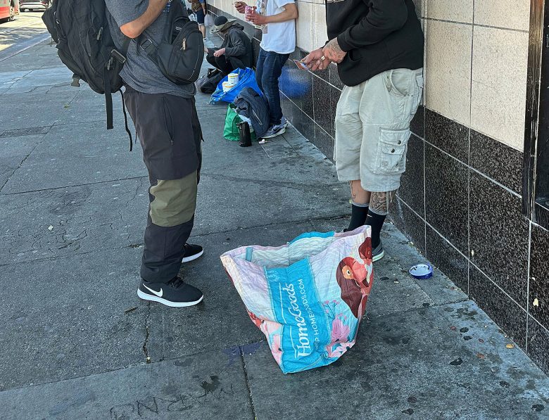 Two people stand on a sidewalk near a wall with a large, partially open shopping bag on the ground. Other individuals and belongings are visible in the background.
