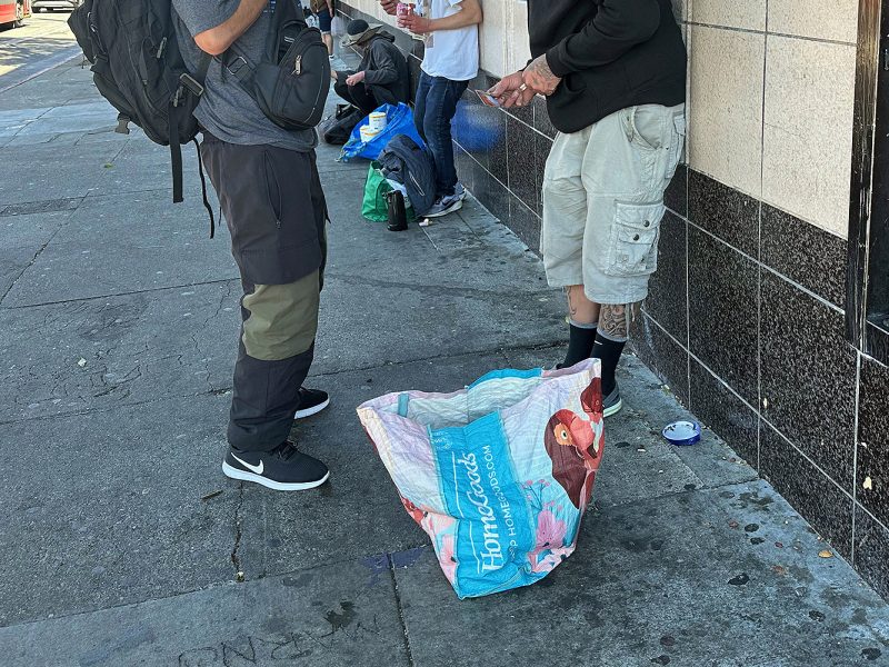 Two people stand on a sidewalk near a wall with a large, partially open shopping bag on the ground. Other individuals and belongings are visible in the background.