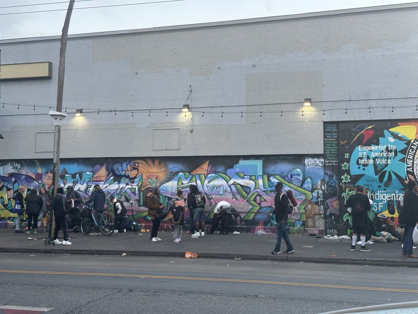 A group of people stand and sit along a sidewalk in front of a building with colorful graffiti and murals on the wall.