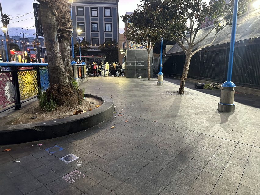 A group of people stands near a building at dusk in an urban plaza with trees, a painted fence, and scattered litter on the ground.