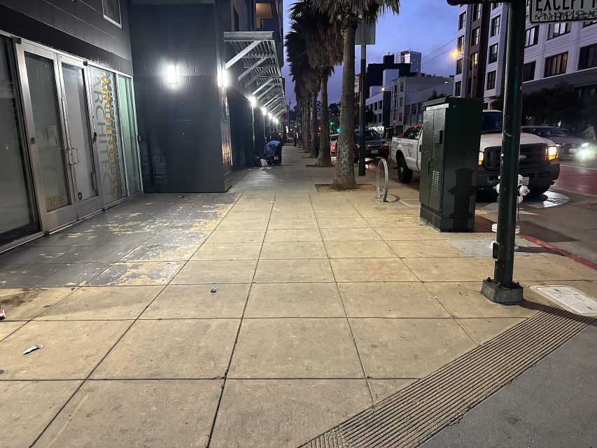 A mostly empty city sidewalk at dusk with a few scattered pieces of litter, palm trees, parked cars, and buildings under streetlights.