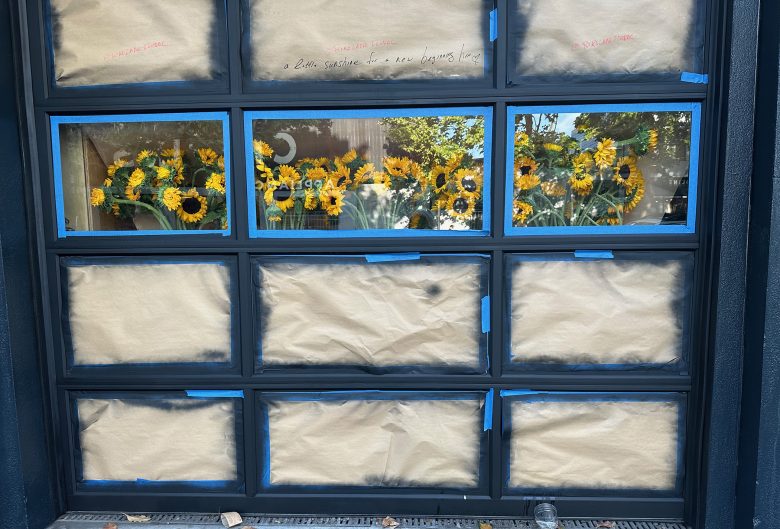 A glass garage door with most panels covered by brown paper, leaving the top row uncovered and revealing vases of sunflowers inside.