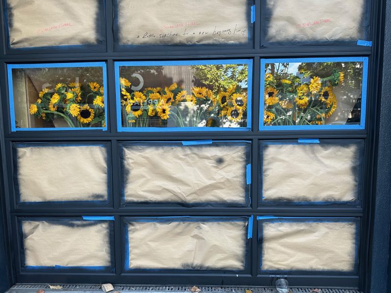 A glass garage door with most panels covered by brown paper, leaving the top row uncovered and revealing vases of sunflowers inside.