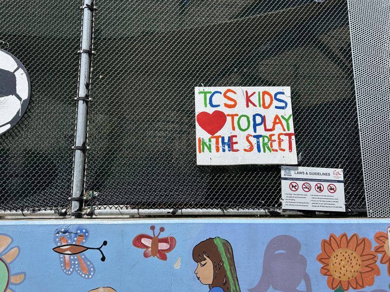 A colorful sign on a chain-link fence in Elm Alley reads “TCS KIDS ❤️ TO PLAY IN THE STREET” above a mural with flowers, a butterfly, and a child’s profile.