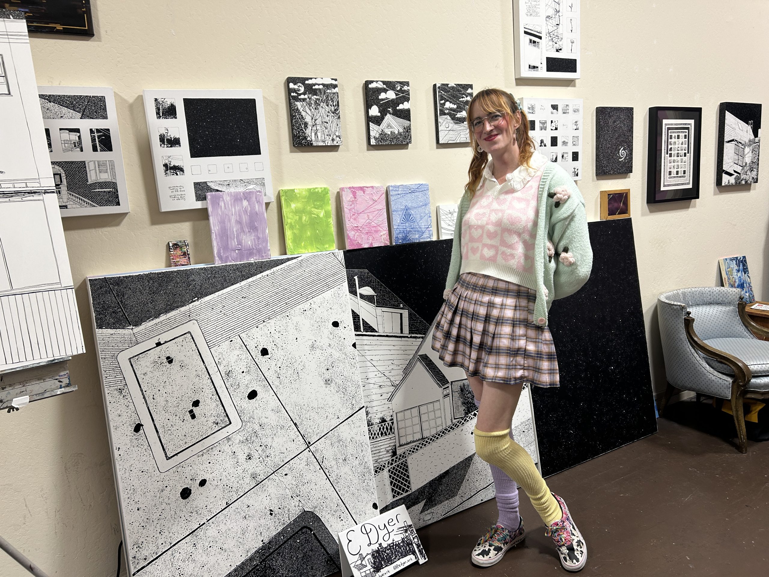 A woman with pigtails stands in front of a wall displaying monochrome and pastel artwork at an art walk, wearing a pastel sweater, plaid skirt, and mismatched knee-high socks.
