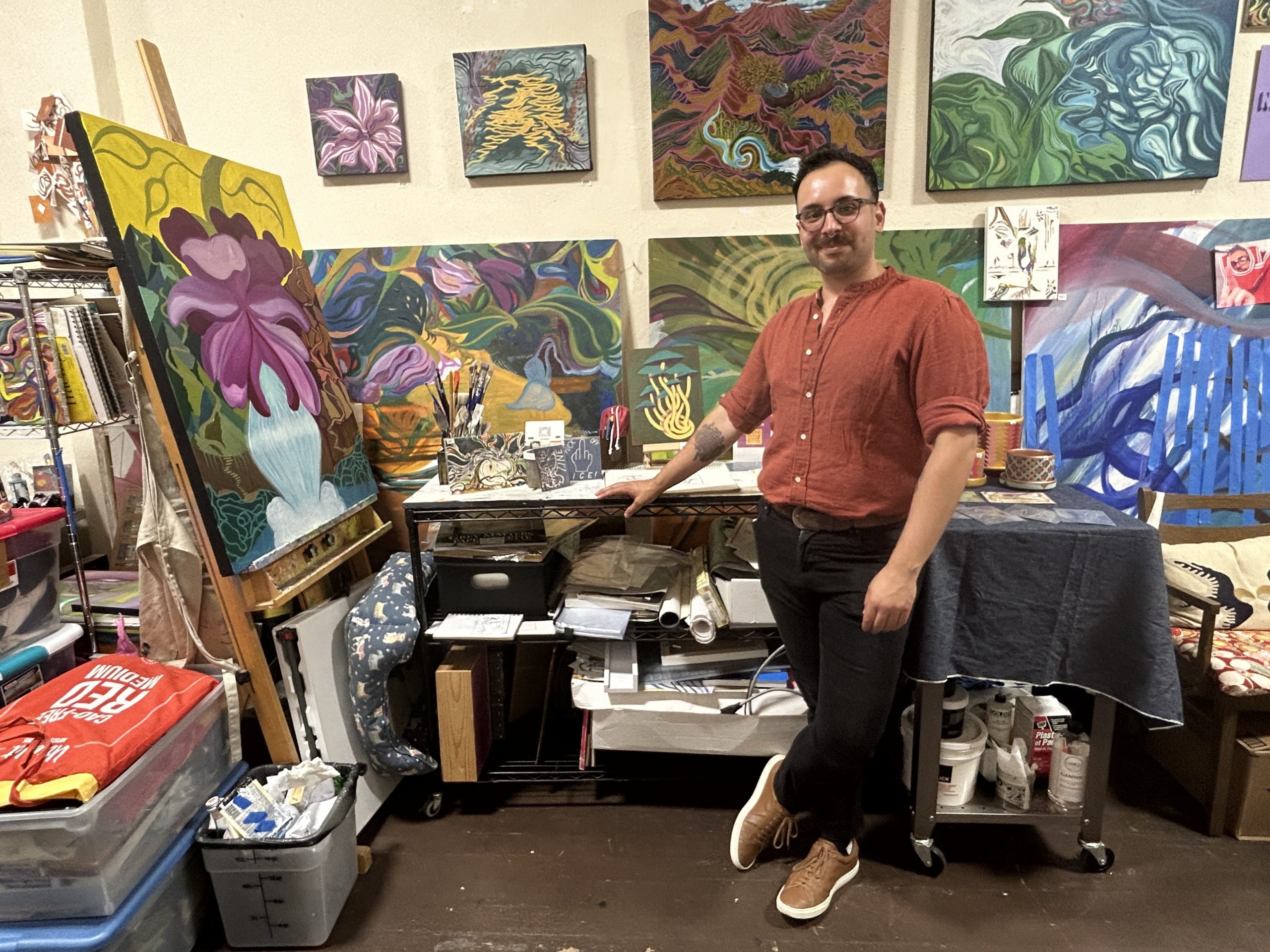 A person in a red shirt stands in an art studio surrounded by colorful abstract paintings, art supplies, and storage bins, preparing for the upcoming art walk.