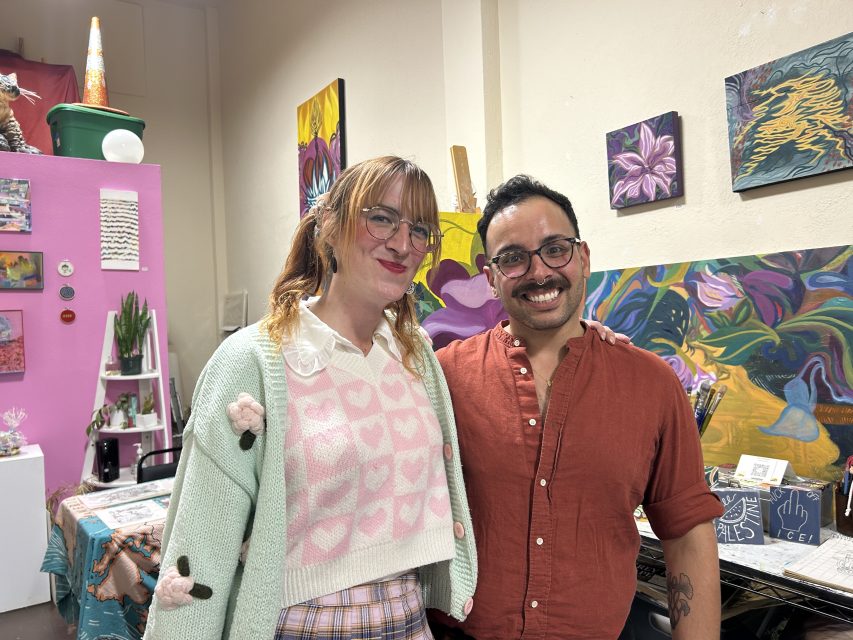 Two people standing side by side and smiling in an art studio with colorful paintings and plants in the background.