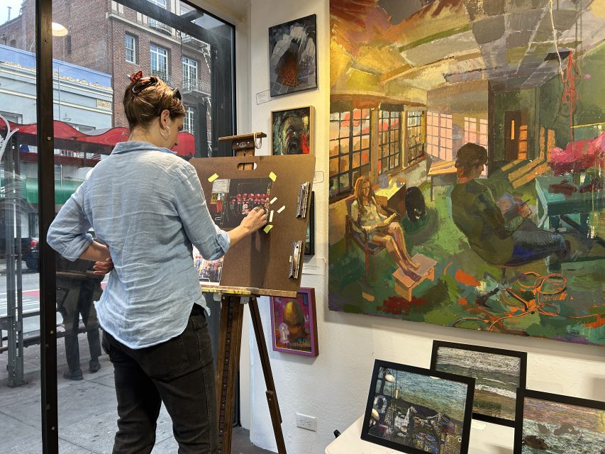 An artist stands at an easel, drawing with pastels in a gallery space filled with colorful paintings and framed art near a large window.
