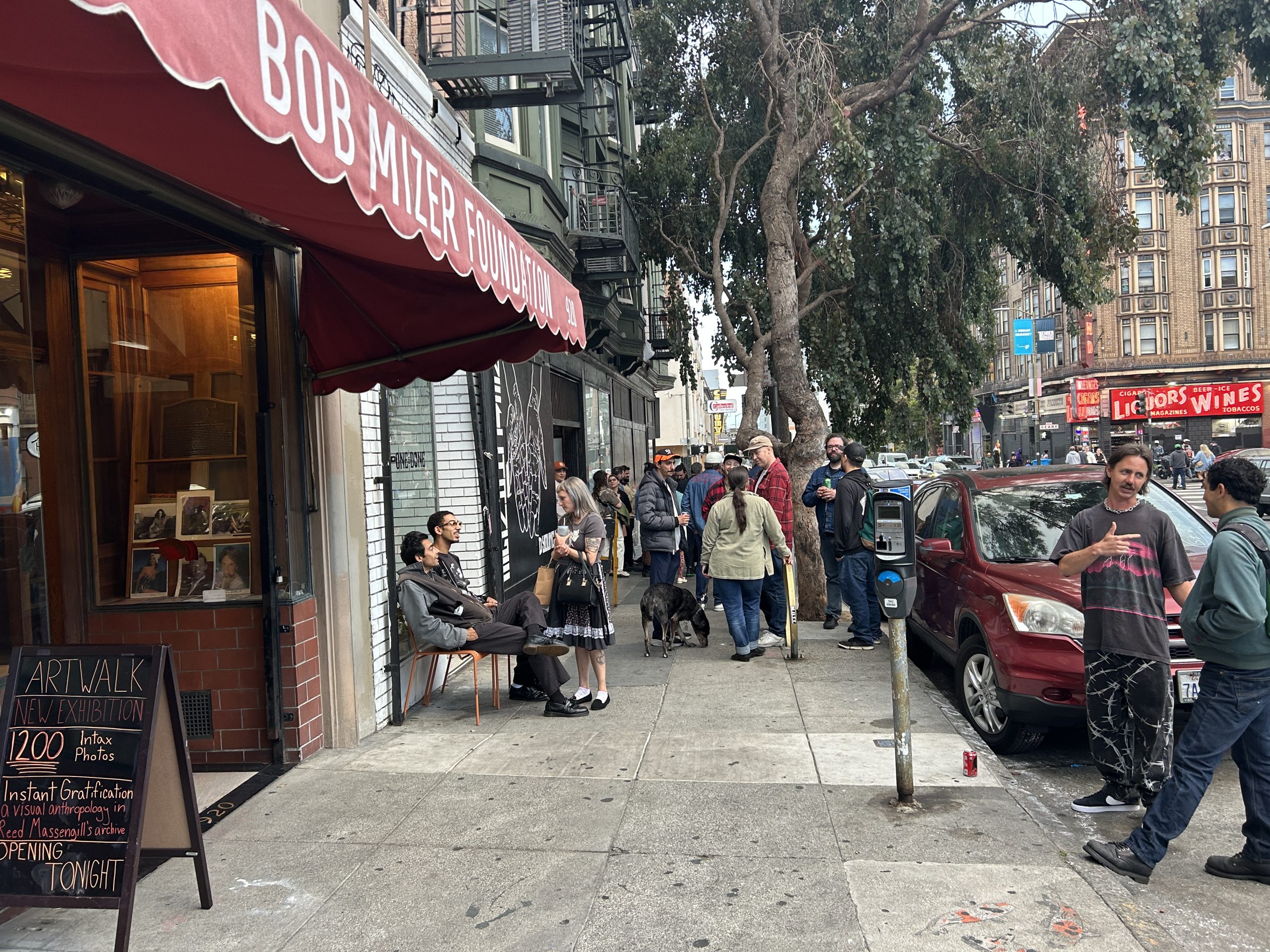 A group of people gather outside the Bob Mizer Foundation on a city sidewalk near an art walk event sign, with parked cars and trees lining the street.