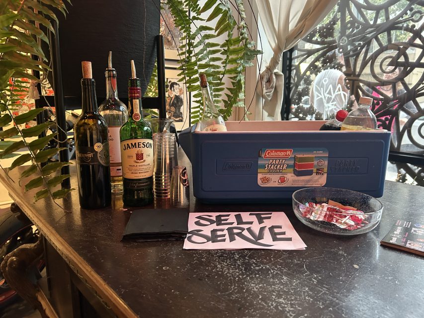 A table displays liquor bottles, plastic cups, a Coleman ice chest, a bowl of condiment packets, and a sign reading "SELF SERVE.