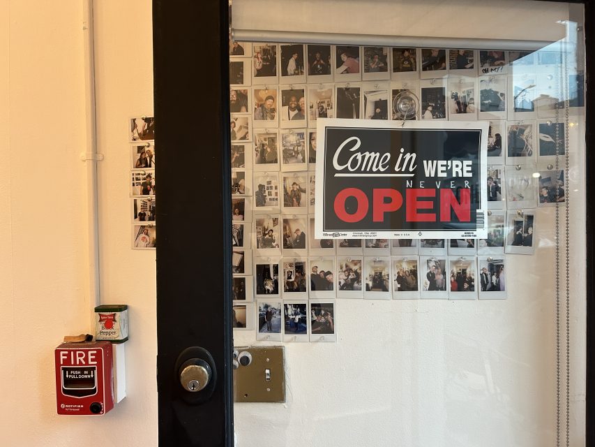 Glass door with a humorous sign reading "Come in We're Never Open," surrounded by Polaroid photos; a fire alarm and can are mounted on the wall nearby.