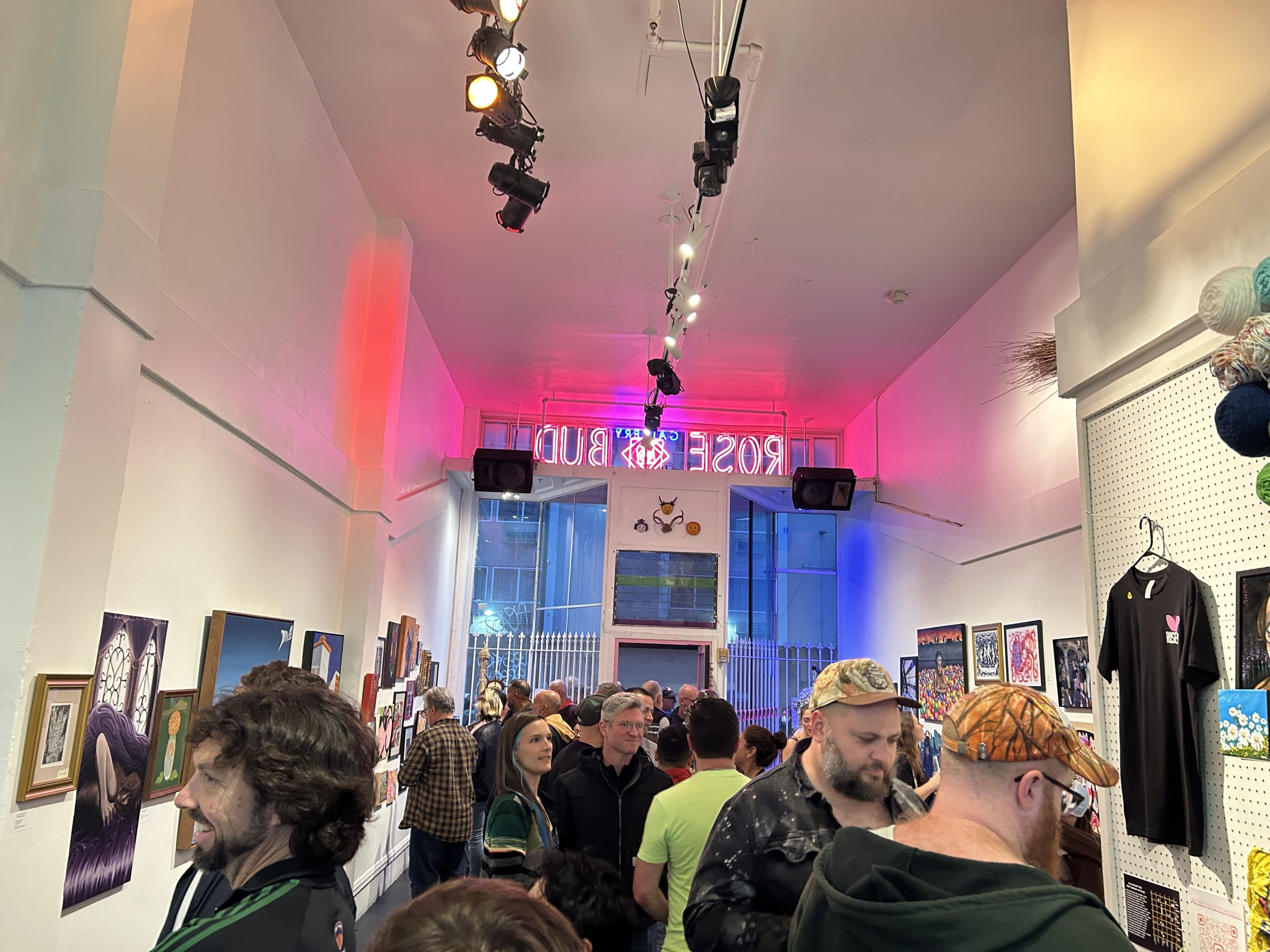 A crowd of people gathers inside an art gallery with colorful lighting and artworks displayed on the walls; a neon "ROSE BUD" sign is visible in the background.