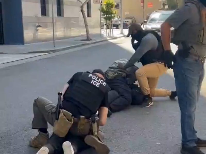 Two ICE officers restrain two individuals on the ground in the middle of a city street while a third officer stands nearby.