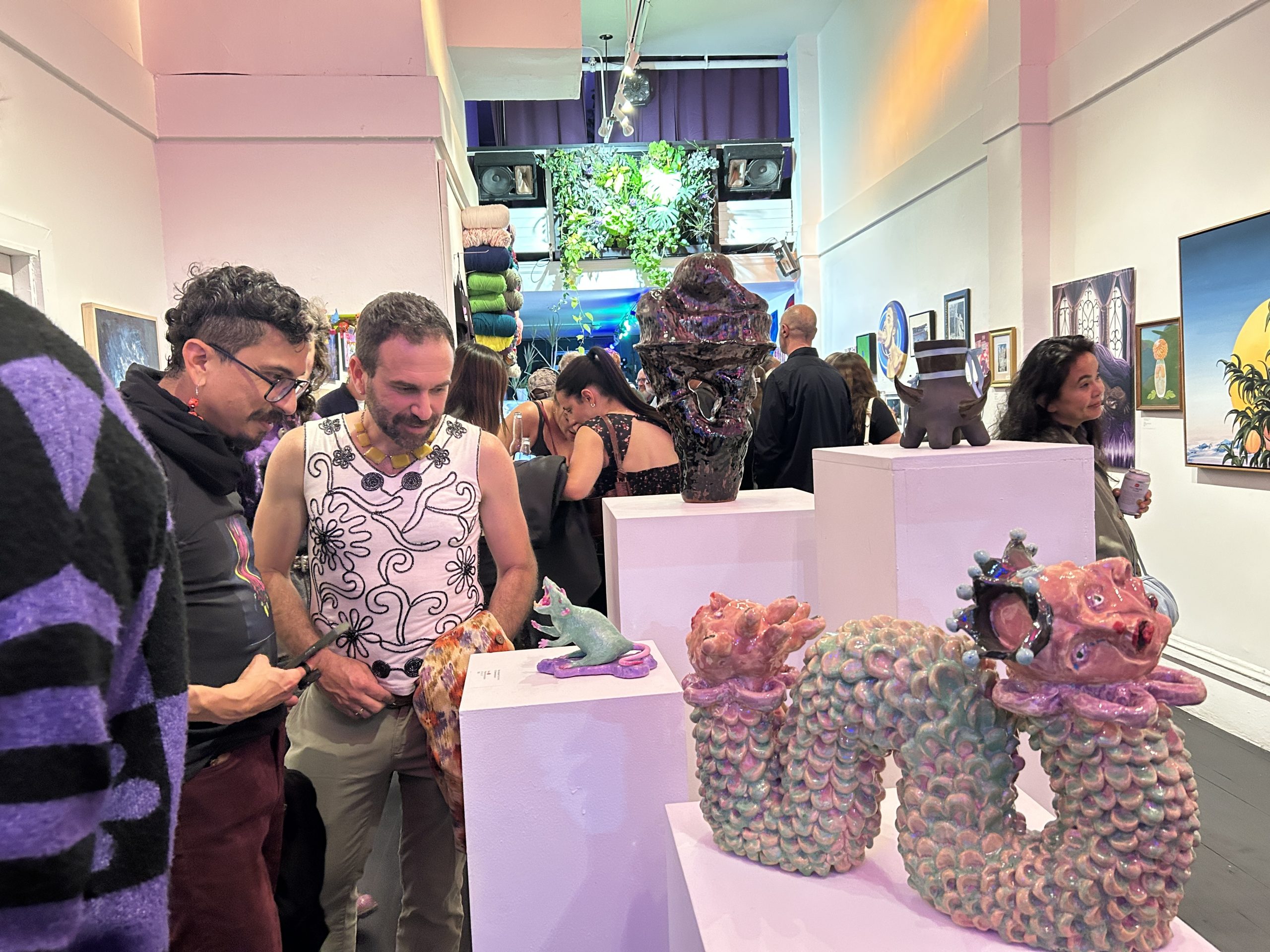 People enjoy an art walk, viewing colorful ceramic sculptures and artwork displayed on white pedestals and walls in a brightly lit art gallery.