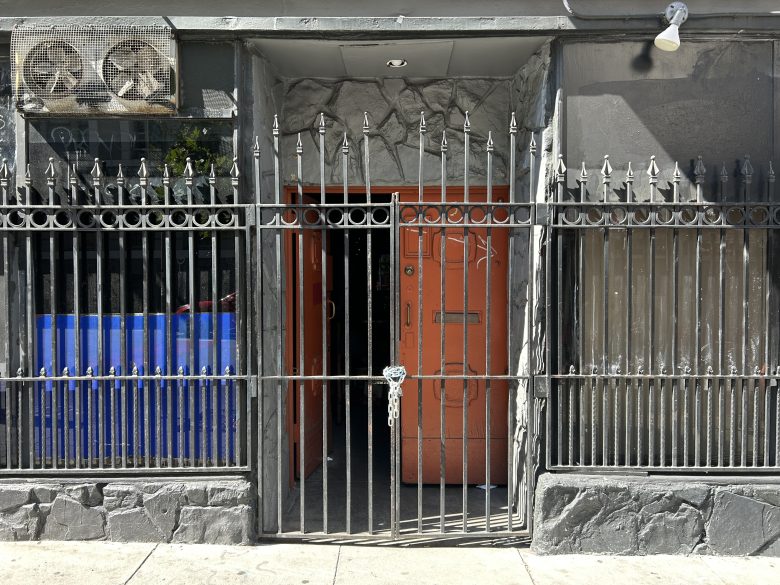 An iron fence with a locked gate stands in front of a building entrance with an open orange door, barred windows, and air conditioning units above.