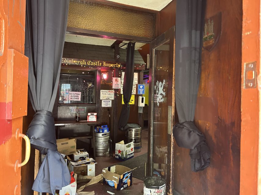 Open doorway to a dimly lit bar with black curtains, beer kegs, boxes, and various signs visible inside; the floor appears cluttered with debris.