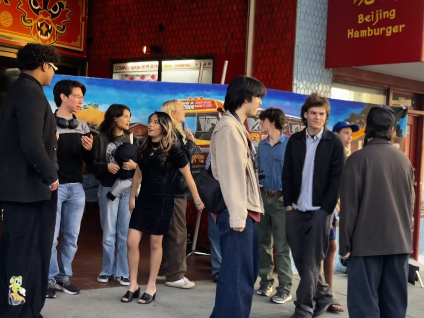 A group of young people stand and converse on a sidewalk outside a restaurant called Beijing Hamburger.