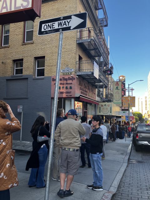 People stand in a long line on a city sidewalk near a one-way street sign, with storefronts and signs visible along the brick building.