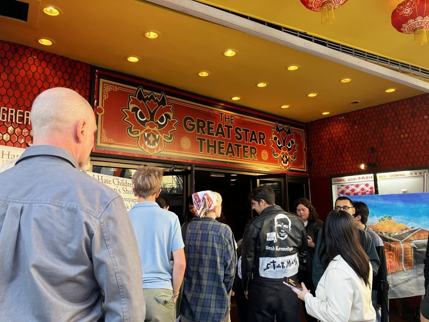 A group of people stands in line outside the entrance to The Great Star Theater, which has a red sign with a stylized mask logo above the door.