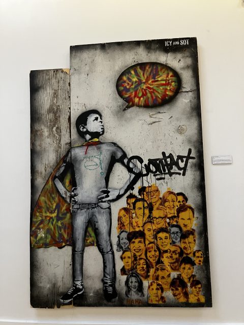 A mixed media artwork shows a boy in a cape with a speech bubble and the word “Sorry,” standing above a crowd of illustrated faces on a wooden panel.