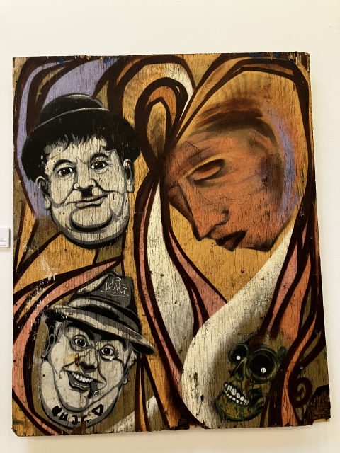 A painting on wood featuring abstract swirls, a large melancholy face in profile, two cartoonish male faces, and a green skull.