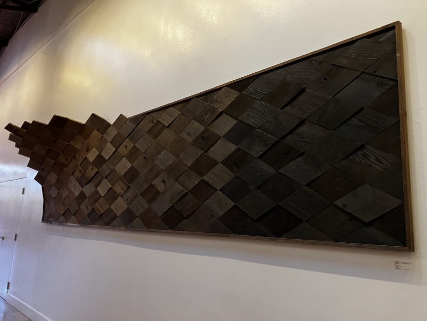 A wall-mounted artwork composed of dark wooden tiles arranged in a geometric, gradually undulating pattern within a rectangular frame.