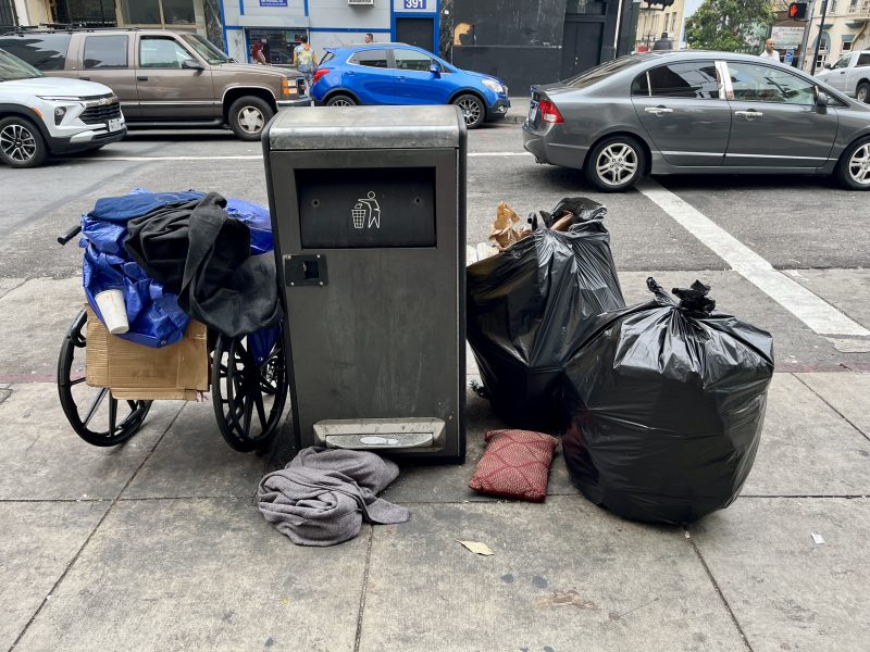A trash bin on a city sidewalk is surrounded by black garbage bags, a cart with a blue tarp, cardboard, and scattered clothing.