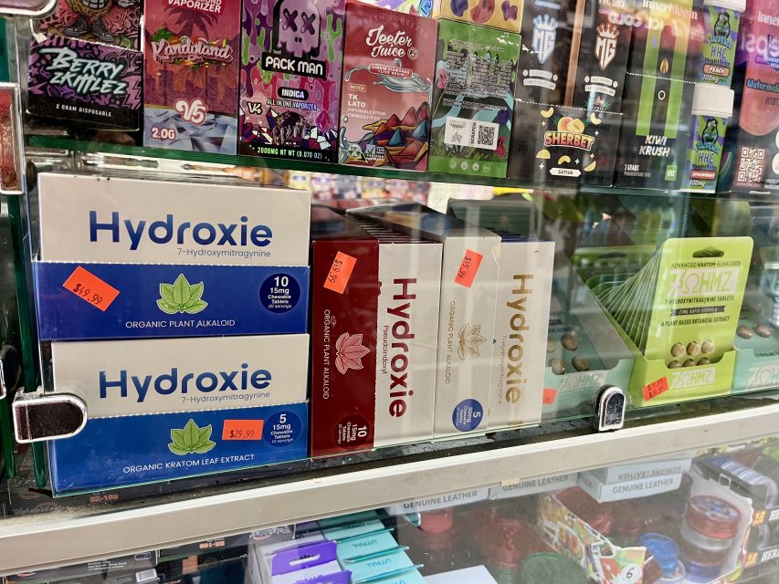 Boxes of Hydroxie kratom leaf extract and other supplements are displayed on a glass shelf in a store, with price tags visible on the packaging.