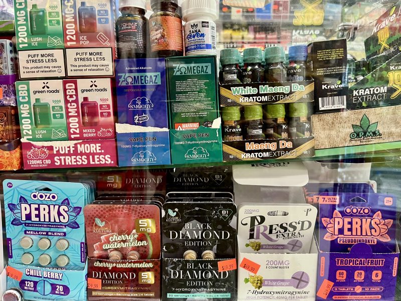 A display case shows various CBD, kratom, and vape products, including gummies, drinks, and extracts, with colorful packaging and visible labels and prices.