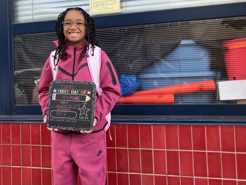 A girl in a pink tracksuit smiles and holds a chalkboard sign reading "First Day of 5th Grade" while standing in front of a building with red tiles and a window.