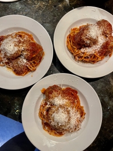 Three white plates of spaghetti with tomato sauce, meatballs, and grated cheese on top, arranged on a dark surface.