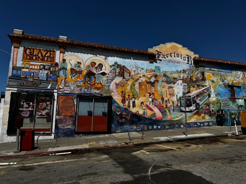 A colorful mural covers the side of a building, depicting scenes of community life, culture, and a donut shop under a clear blue sky.