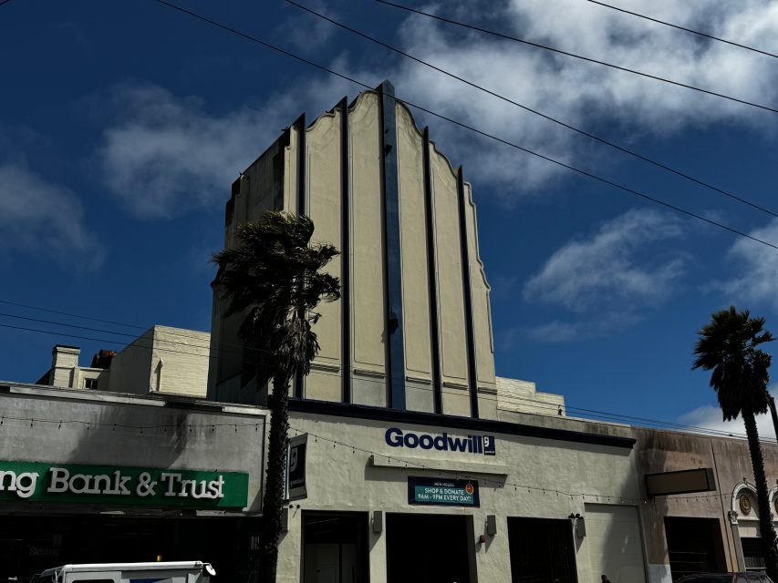 A beige Art Deco building with a Goodwill store on the ground floor stands between two palm trees under a partly cloudy sky.