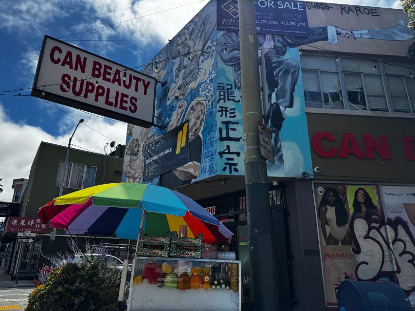 Street view of a beauty supply store with a colorful fruit stand, a large mural, and signage in both English and Chinese.