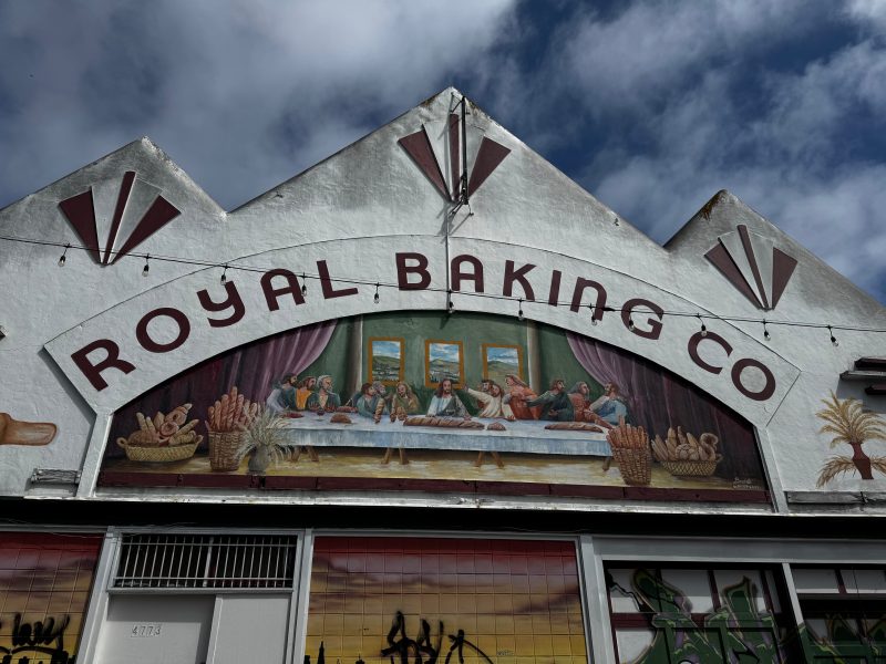 A bakery storefront with a painted mural resembling The Last Supper, featuring bread and pastries, under the sign "Royal Baking Co" against a partly cloudy sky.