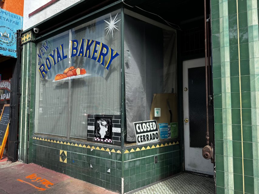 A bakery storefront with "New Royal Bakery" painted on the window, a “Closed/Cerrado” sign, and green tiled walls. The shop appears to be currently closed.