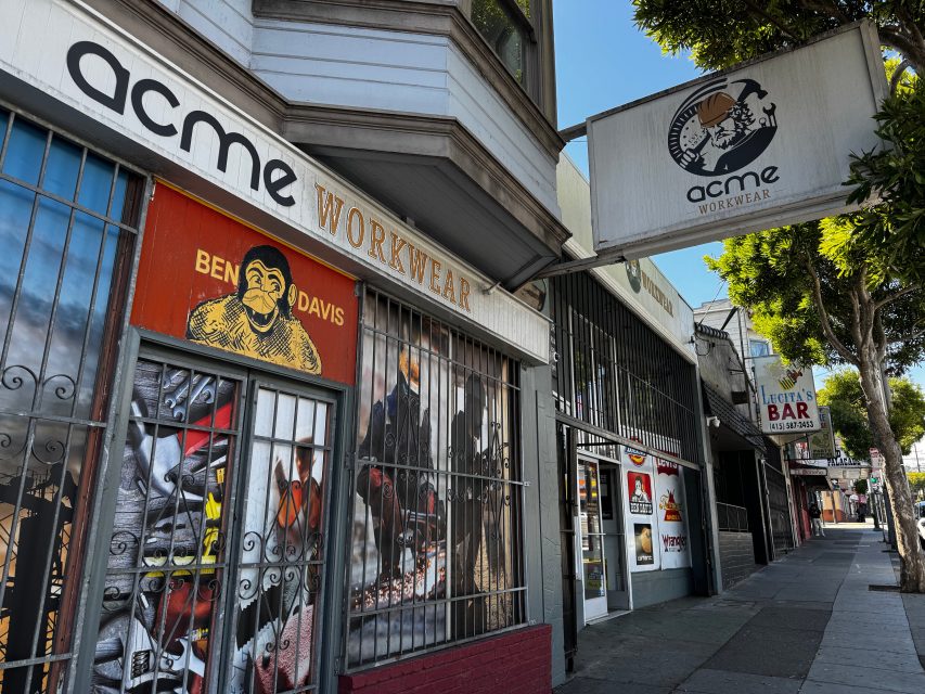 Street view of storefronts including Acme Workwear and Ben Davis, with barred windows and signage, on a sunny day with tree-lined sidewalk.
