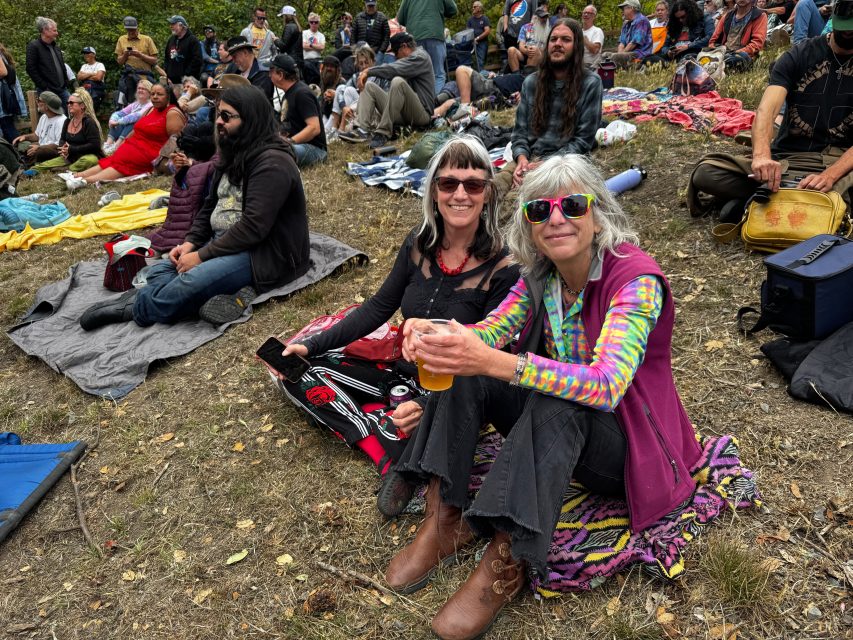 A group of people sits on blankets and the ground at an outdoor event; two women in the foreground smile at the camera, one holding a drink.