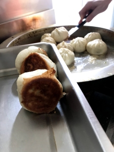A person uses a spatula to cook and transfer Chinese pan-fried buns with golden brown bottoms onto a metal tray.