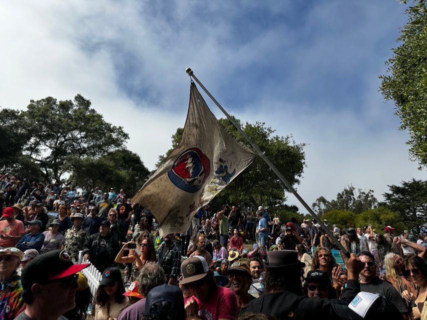 A large crowd gathered outdoors under a cloudy sky, with a worn Grateful Dead flag raised in the center among trees.