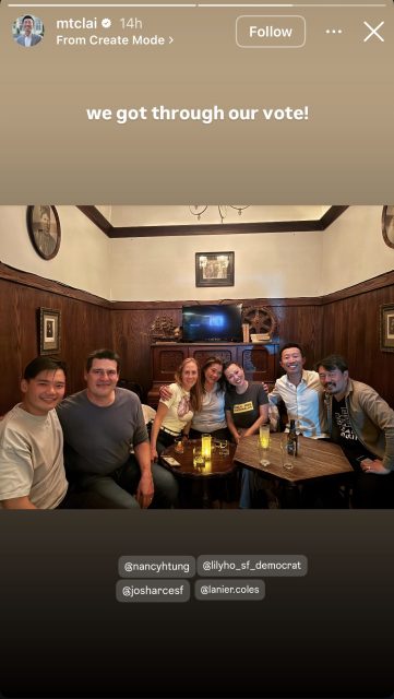 Seven people are sitting together around a small table in a dimly lit paneled room, smiling at the camera with drinks in front of them.