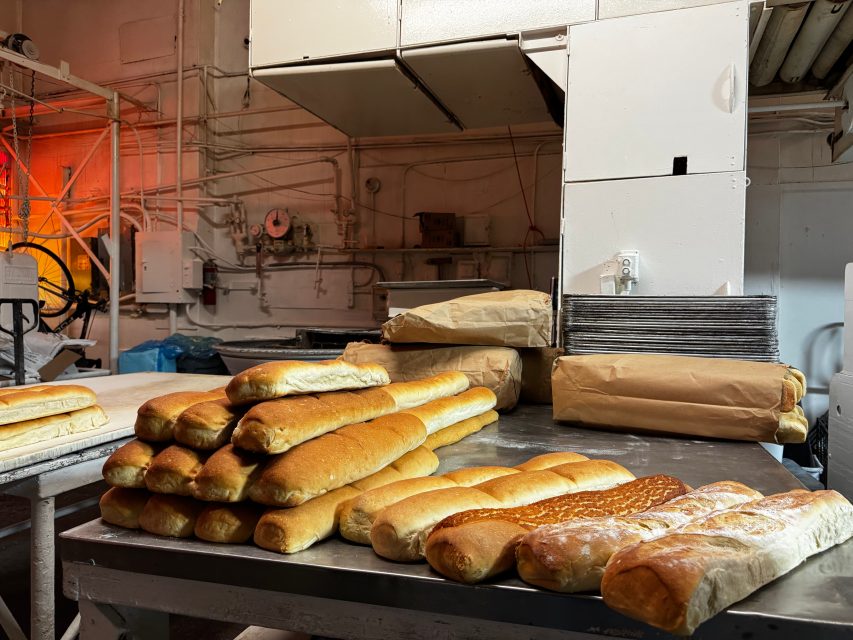 Stacks of fresh baguettes and loaves are arranged on a metal table in an industrial bakery with machinery and pipes in the background.