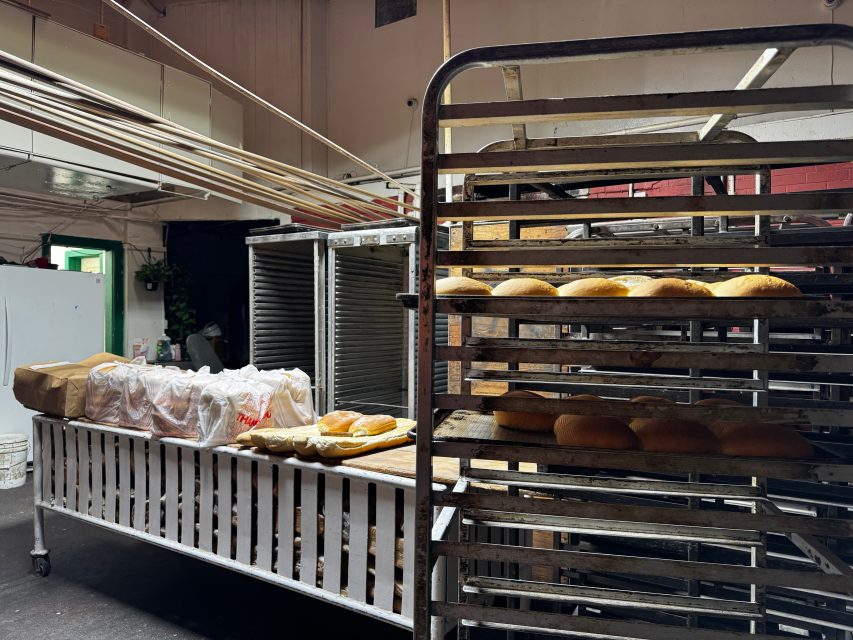 Racks of freshly baked bread loaves and packaged bread are arranged on metal shelves and a striped table in a bakery setting.