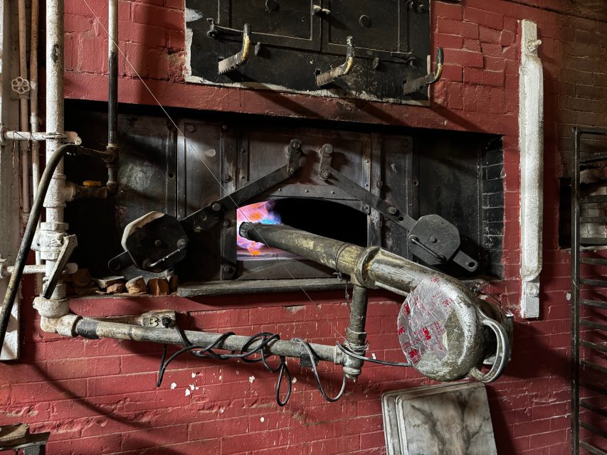 Industrial furnace with metal pipes and a burner flame visible through an open hatch set in a red brick wall.
