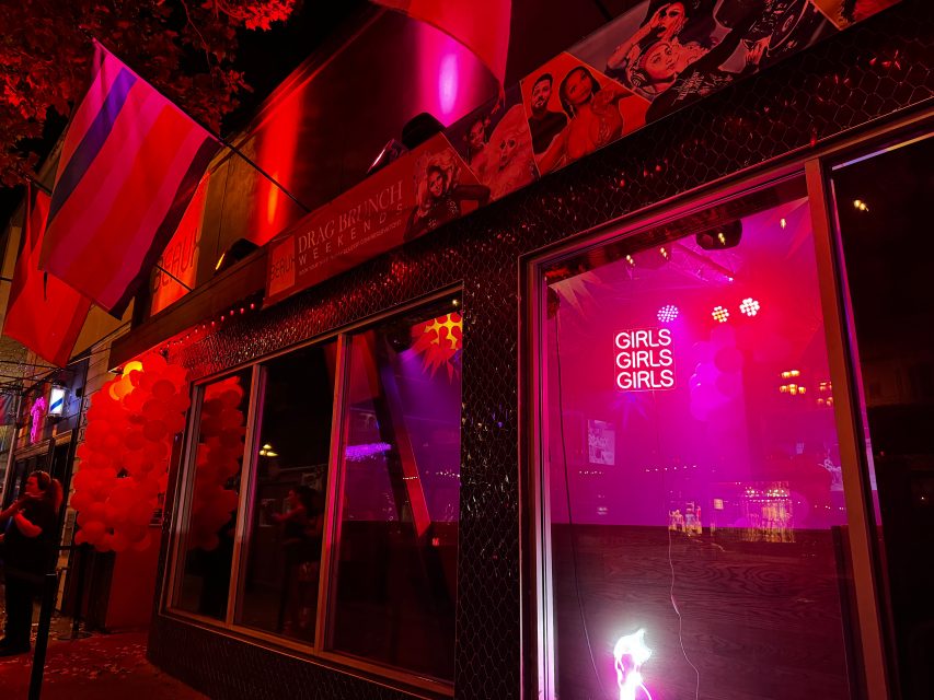 A bar exterior at night with neon "GIRLS GIRLS GIRLS" sign, pink lighting, pride flags, and balloons, creating a festive lesbian-friendly atmosphere.