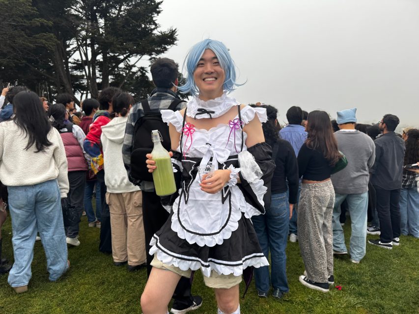 Person wearing a blue wig and black-and-white maid costume holding a green drink, standing outdoors in front of a crowd on grass.