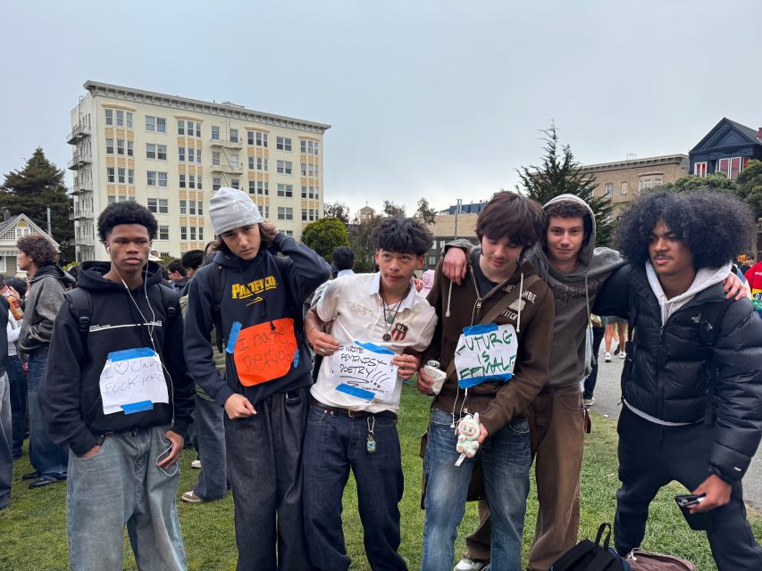 Six teenagers stand together outside on grass, each wearing handmade signs with messages; an apartment building and other people are visible in the background.