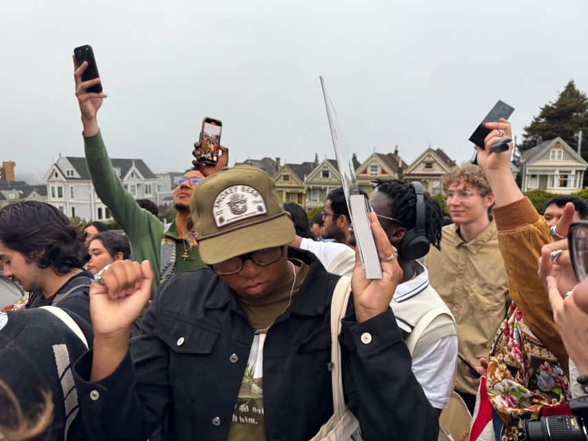 A group of people outdoors, some raising their arms and holding phones, while others wear headphones. Victorian-style houses are visible in the background.